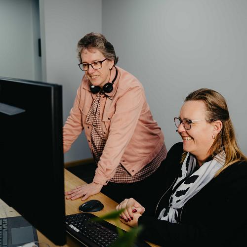 Twee collega’s kijken samen glimlachend naar een computerscherm in een kantoorruimte, terwijl één van hen aanwijzingen geeft.