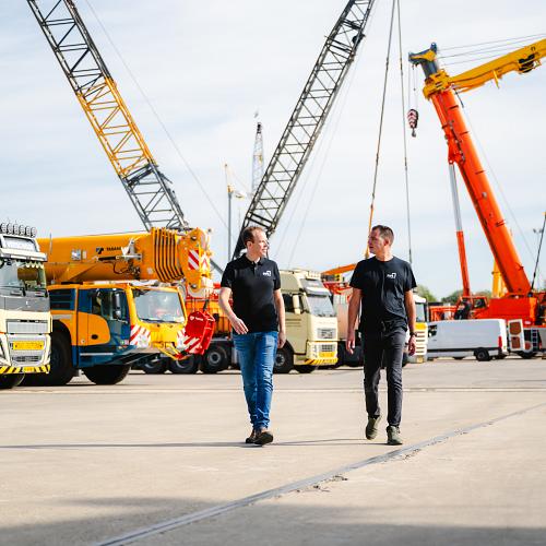 Twee PWA-medewerkers lopen over het terrein van een kraan- en transportbedrijf, tussen indrukwekkende voertuigen en hijskranen.