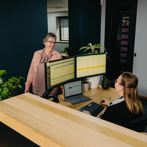 Twee PWA-medewerkers overleggen bij een werkplek met meerdere schermen, in een modern kantoor met planten en een open sfeer.