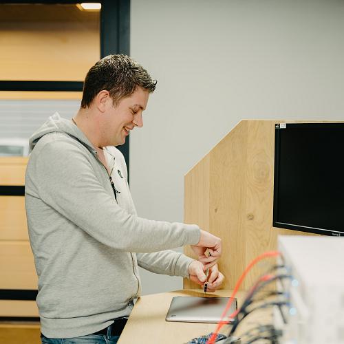 Lachende man staat achter een bureau en werkt op een computer in een moderne kantooromgeving, bezig met een schroevendraaier en een laptop.
