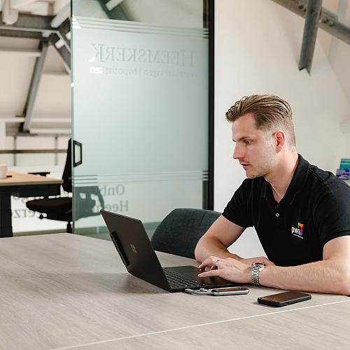 Lachende man staat achter een bureau en werkt op een computer in een moderne kantooromgeving, bezig met een schroevendraaier en een laptop.