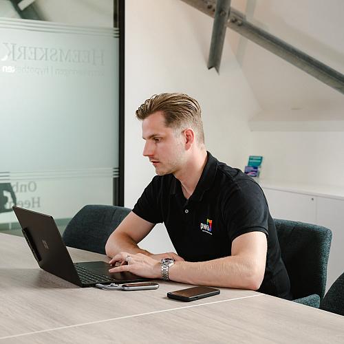 Lachende man staat achter een bureau en werkt op een computer in een moderne kantooromgeving, bezig met een schroevendraaier en een laptop.