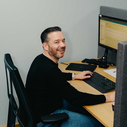 Lachende man zit achter een bureau en werkt op een computer in een moderne kantooromgeving met grijze schermwanden.