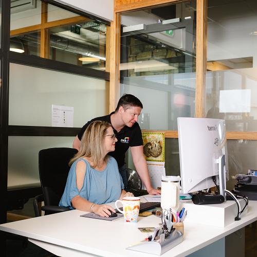 IT consultant helpt klant achter een bureau; een vrouw zit achter de computer terwijl een man in PWA-bedrijfskleding meekijkt op het scherm in een modern kantoor.