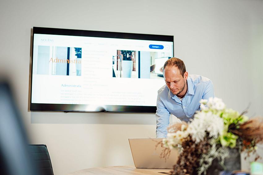 Man werkt geconcentreerd op zijn laptop tijdens een meeting, met een grote presentatiescherm op de achtergrond.
