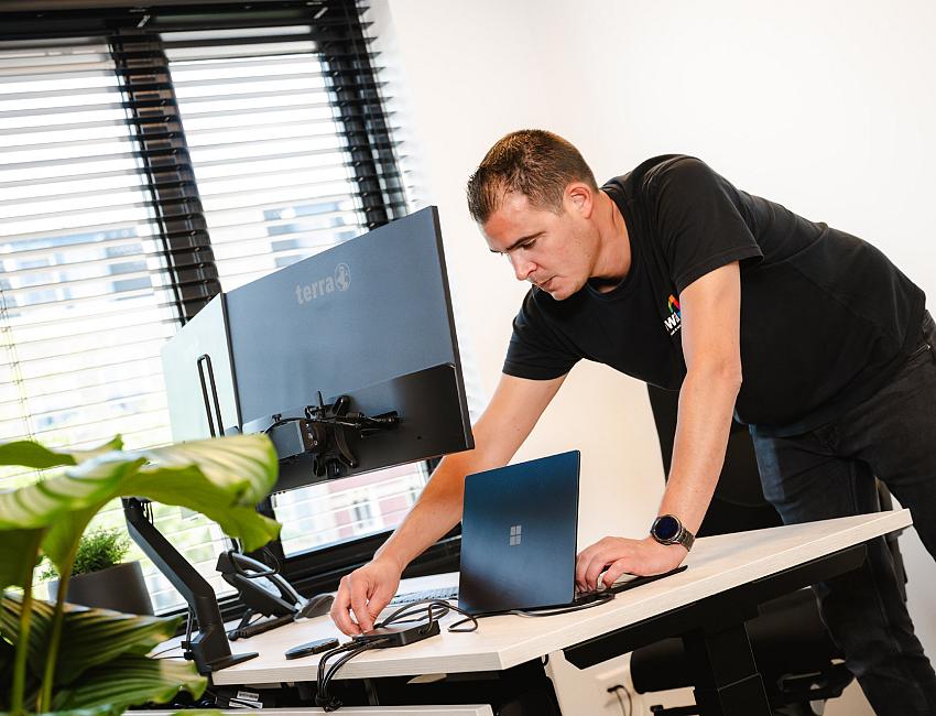 PWA-medewerker sluit een laptop aan op een werkplek met dubbele monitoren in een modern kantoor met veel daglicht en planten.
