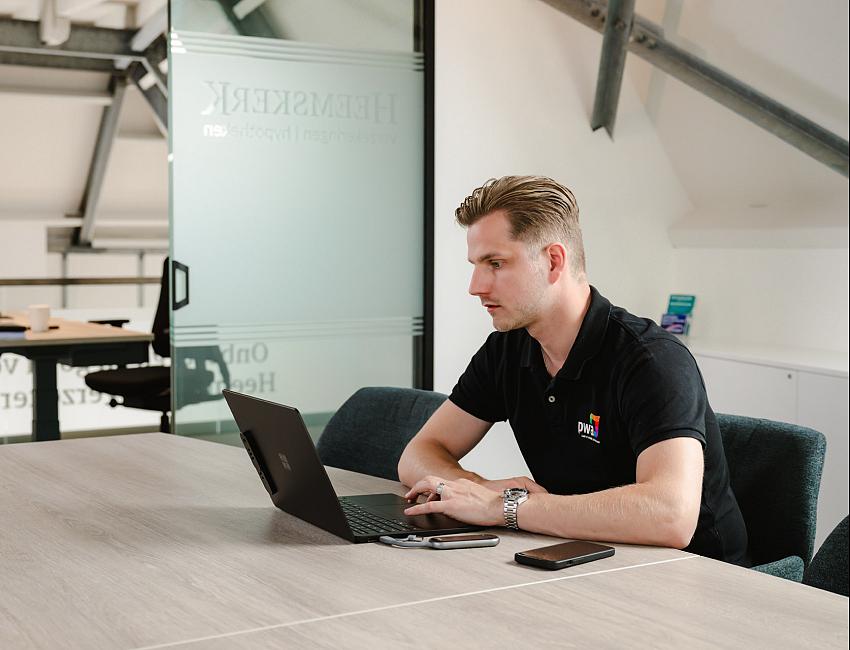 Man in PWA-bedrijfskleding werkt geconcentreerd op een laptop aan een vergadertafel in een modern kantoor met glazen wanden en industriële details.