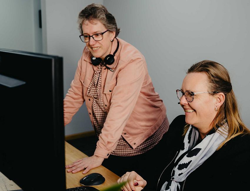 Twee collega’s werken samen achter een computerscherm in een kantoor, waarbij één vrouw staat en meekijkt terwijl de ander glimlachend achter het toetsenbord zit.