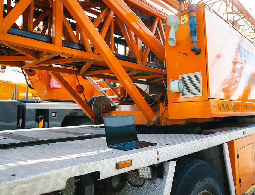 Zwarte, open laptop die midden op het metalen, geribbelde platform van een grote, oranje hijskraanwagen ligt. De feloranje giek en kraanopbouw domineren de achtergrond.