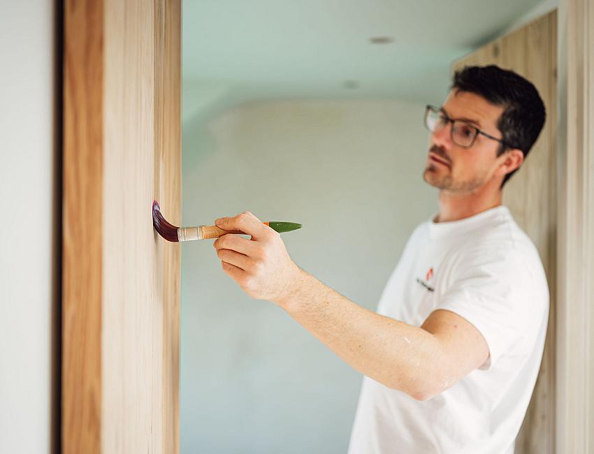 Schilder van Van Kesteren brengt zorgvuldig verf aan op een houten wand in een binnenruimte.