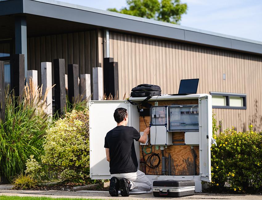Monteur op zijn knieën voor een netwerkkast in een groene omgeving met een stacaravan op de achtergrond. 