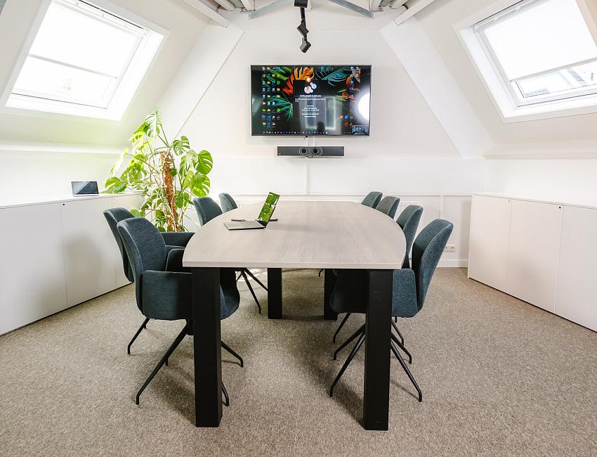 Vergaderruimte bij PWA met ovale tafel, blauwe stoelen en groot scherm voor videomeetings onder een schuin dak met daglicht.