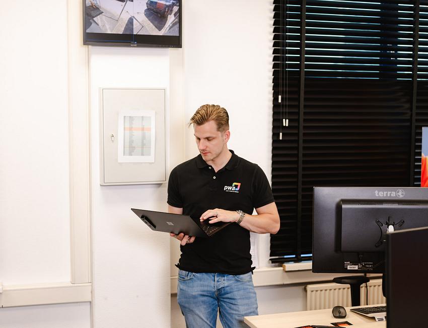 Man in PWA-bedrijfskleding werkt staand op een laptop in een kantoorruimte met beeldschermen en gesloten jaloezieën.