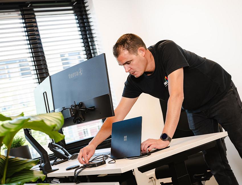 PWA-medewerker stelt een werkplek in en sluit apparatuur aan op een bureau met beeldscherm en laptop.