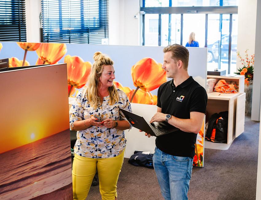 Man en vrouw praten lachend met elkaar in een kleurrijke kantooromgeving met tulpenfoto’s, terwijl de man een laptop vasthoudt.