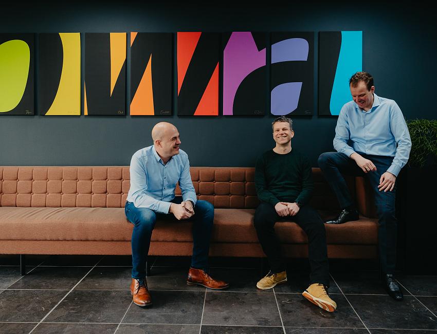 Drie mannen zitten en staan lachend op en rond een bank in een moderne kantoorruimte, met een kleurrijk kunstwerk aan de muur op de achtergrond.