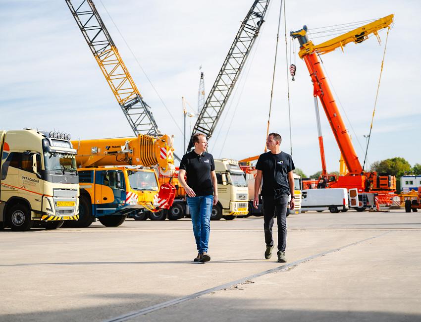 Twee mannen in PWA-kleding lopen over een bedrijfsterrein met meerdere hijskranen en vrachtwagens, onder een heldere lucht.