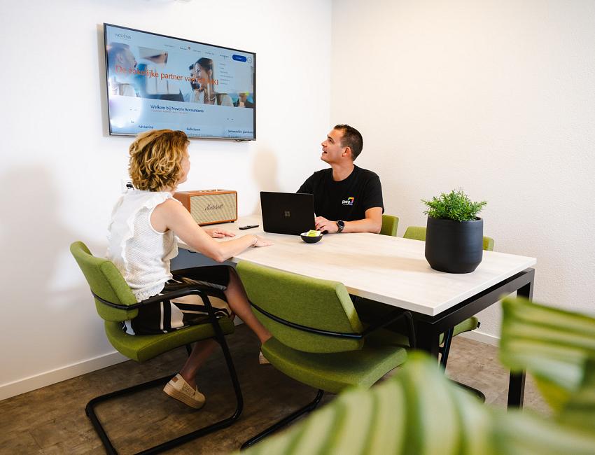 Een klant met een collega in een moderne vergaderruimte, zittend aan een tafel met groene stoelen, in gesprek terwijl een presentatie op het scherm aan de muur zichtbaar is.