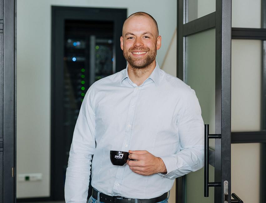 Een glimlachende medewerker in een lichtblauw overhemd staat met een kop koffie in de deuropening van een moderne kantoorruimte.