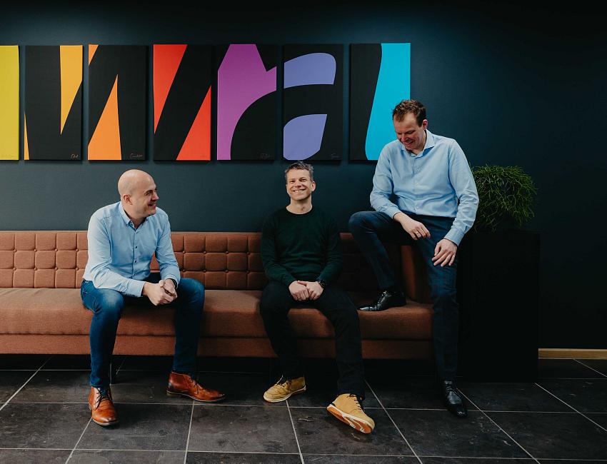 Drie mannen zitten en staan lachend op en rond een bank in een moderne kantoorruimte, met een kleurrijk kunstwerk aan de muur op de achtergrond.