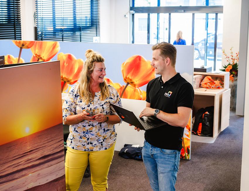 Man en vrouw praten lachend met elkaar in een kleurrijke kantooromgeving met tulpenfoto’s, terwijl de man een laptop vasthoudt.