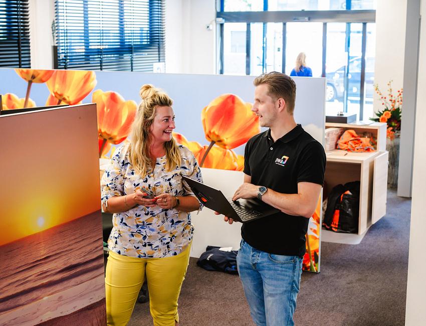 Man en vrouw praten lachend met elkaar in een kleurrijke kantooromgeving met tulpenfoto’s, terwijl de man een laptop vasthoudt.