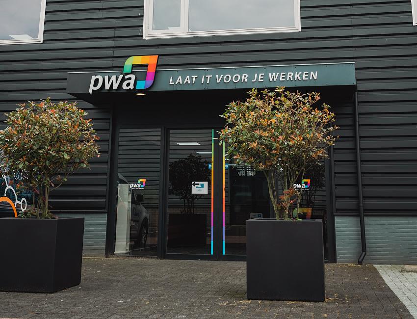 Voorgevel van het PWA-kantoor met logo en slogan boven de entree, geflankeerd dotwee bomen in zwarte plantenbakken.