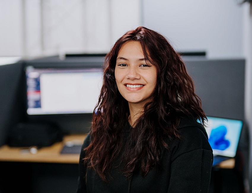 Vrouw met lang donker haar glimlacht terwijl ze achter een werkplek met meerdere schermen zit in een modern kantoor.