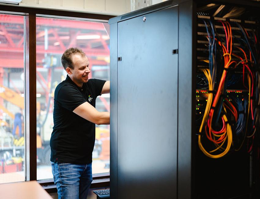 Lachende man in PWA-shirt werkt aan een patchkast vol kleurrijke netwerkkabels, met een industriële werkplaats zichtbaar door het raam op de achtergrond.
