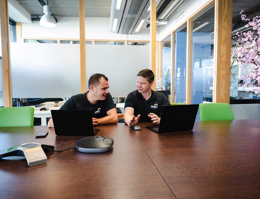 Twee collega’s in PWA-kleding overleggen met hun laptops aan een vergadertafel in een modern kantoor met glazen wanden en bloesemtak.
