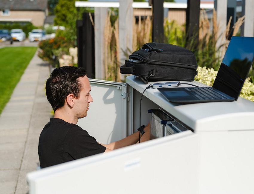 Man werkt aan de binnenkant van een straatkast in een woonwijk, met een open laptop en rugtas bovenop de kast.