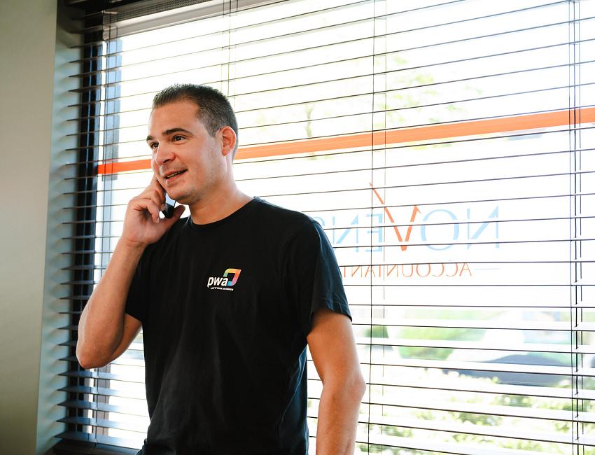 Man in PWA-shirt belt glimlachend bij het raam van een kantoor met jaloezieën en het logo van Novinco op de ruit.