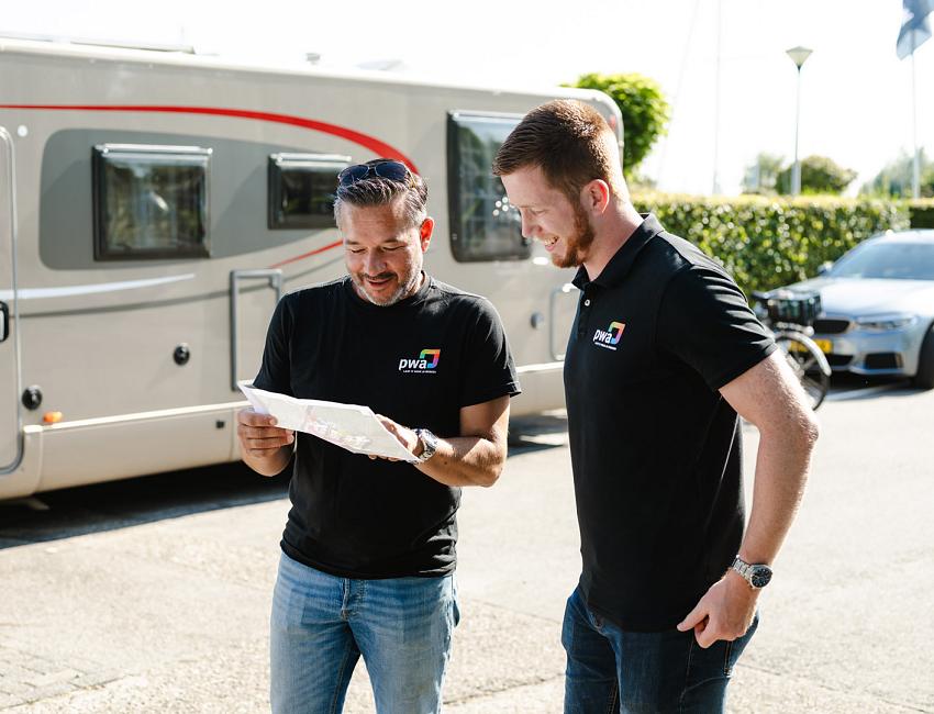 Twee mannen in PWA-shirts bekijken samen een plattegrond of tekening op straat, met een camper en auto op de achtergrond.