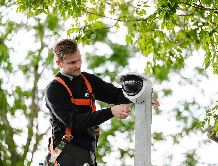 Monteur in veiligheidsharnas installeert een beveiligingscamera aan een paal tussen groene boomtakken.
