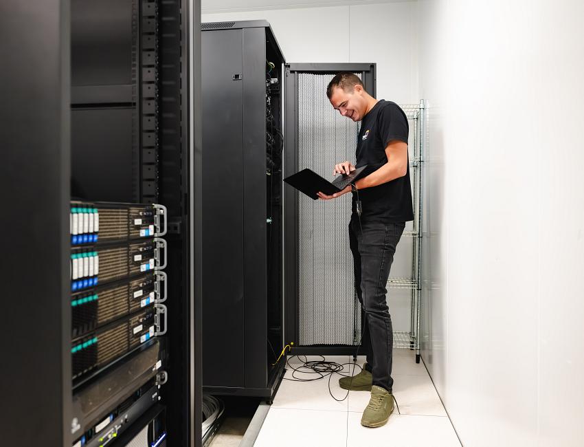 Lachende man staand voor een serverkast met een laptop in zijn hand.