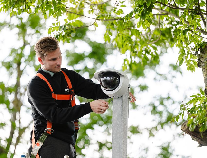 Monteur in veiligheidsharnas installeert een beveiligingscamera aan een paal tussen groene boomtakken.