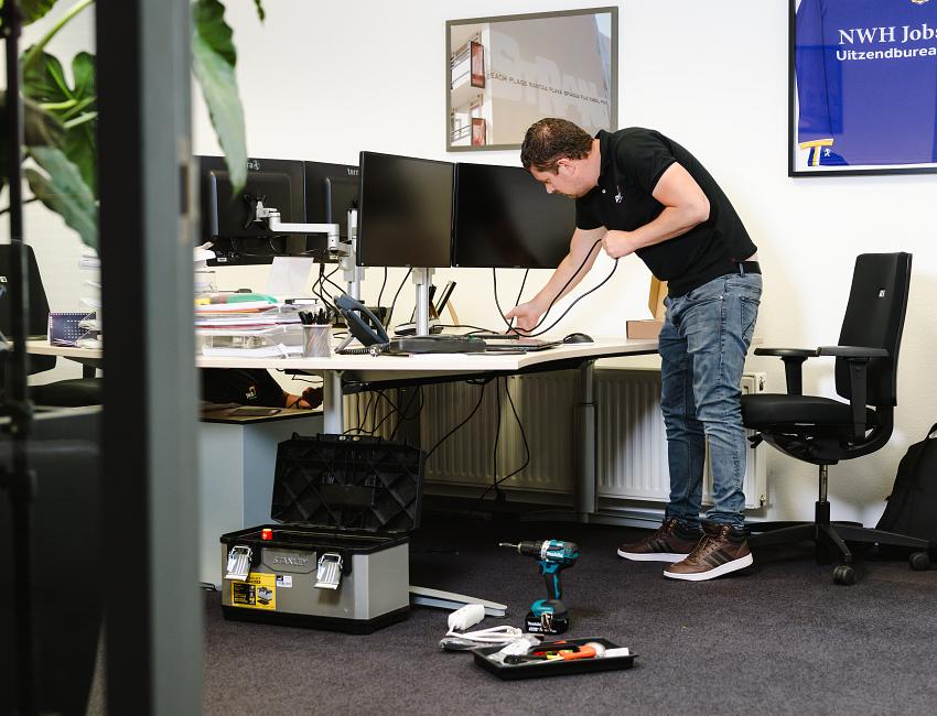 IT-professional richt een werkplek in met meerdere schermen en bekabeling, omringd door gereedschap in een zakelijke kantooromgeving.