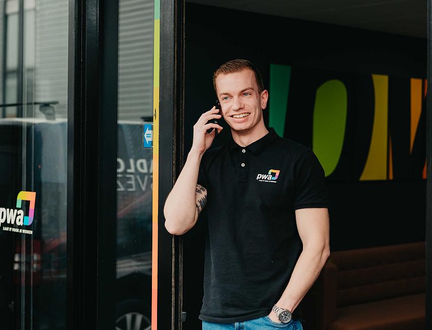 Lachende man in PWA-shirt belt staand bij de ingang van een modern kantoor met kleurrijke wanddecoratie.