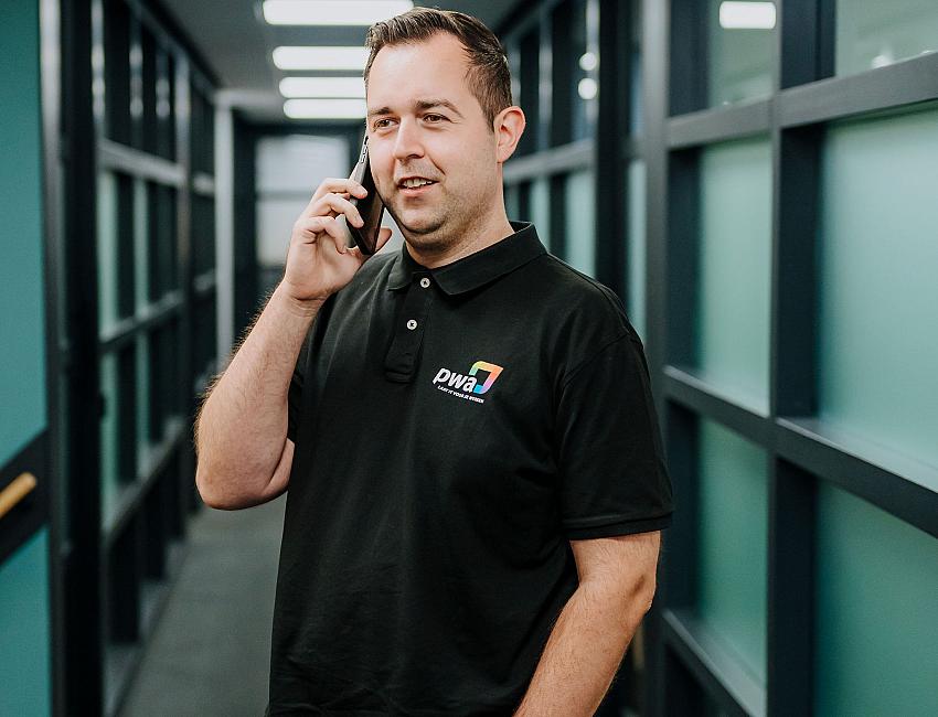 Man in PWA-bedrijfskleding voert een telefoongesprek in een moderne kantoorhal met glazen wanden en tl-verlichting.