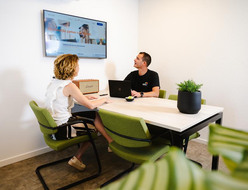 PWA-medewerker in overleg met een klant aan een tafel, met laptops en een presentatie op een scherm aan de muur.