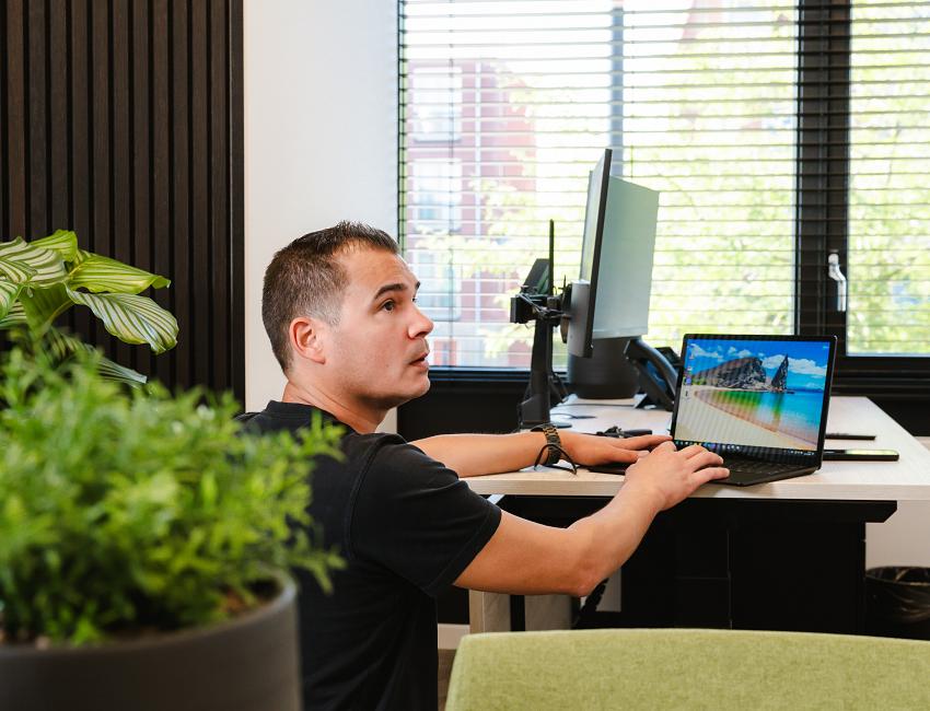 PWA-medewerker richt een werkplek in met laptop en monitor, in een modern kantoor met veel daglicht en groene accenten.