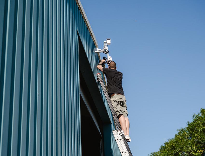Monteur in PWA-bedrijfskleding installeert een beveiligingscamera op de gevel van een blauw bedrijfsgebouw, staand op een ladder onder een heldere blauwe lucht.