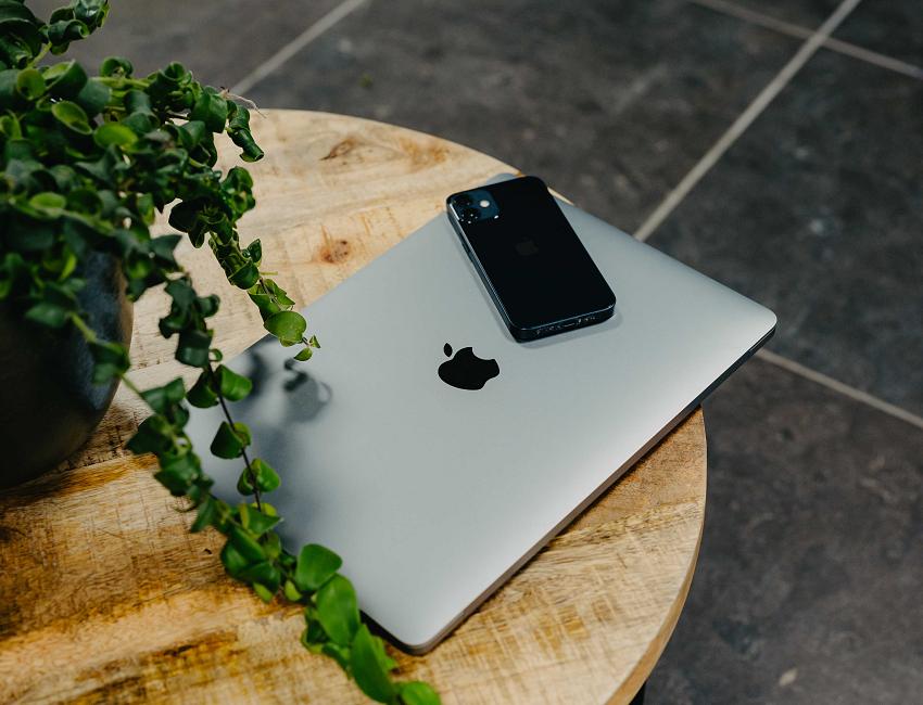 Een gesloten laptop met een smartphone erop ligt op een ronde houten tafel naast een groene plant.
