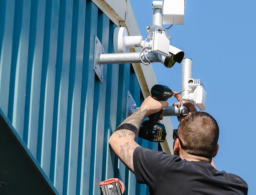 Monteur in veiligheidsharnas installeert een beveiligingscamera aan een paal tussen groene boomtakken.