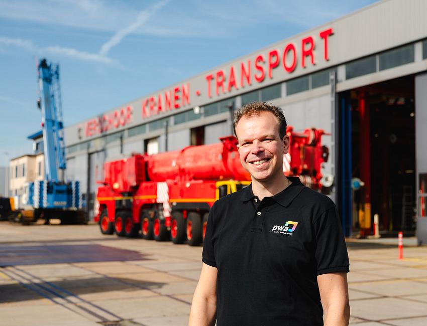 Man met zwart shirt voor loods met rode hijskraanwagen op de achtergond.