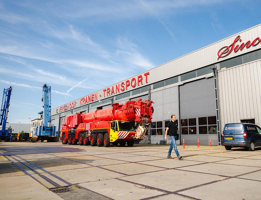 Man met zwart shirt loopt voor loods met rode hijskraanwagen op de achtergond.