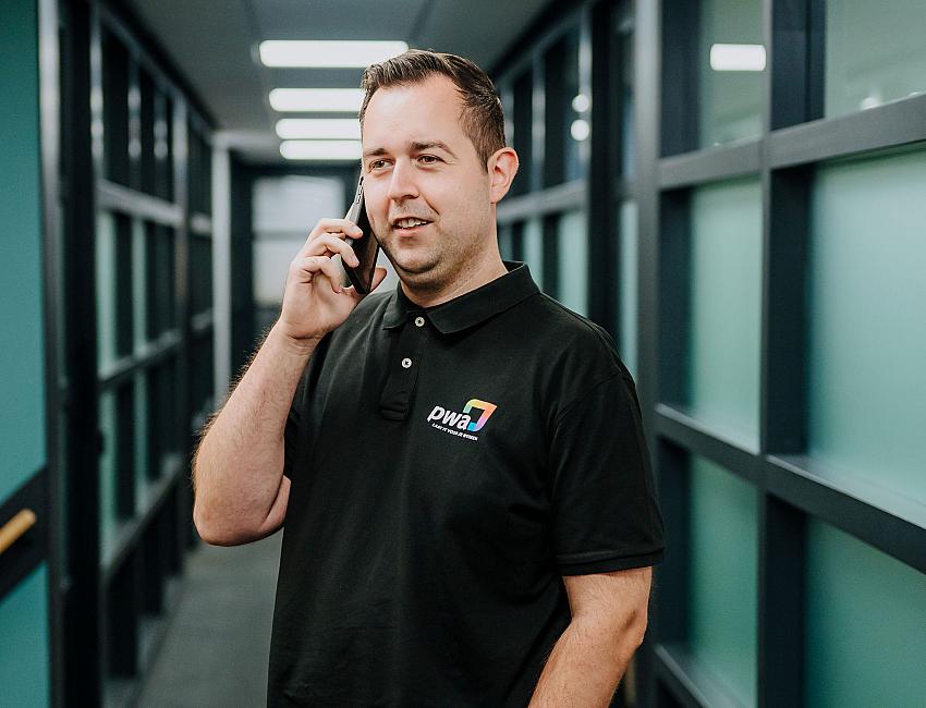 Man in PWA-bedrijfskleding voert een telefoongesprek in een moderne kantoorhal met glazen wanden en tl-verlichting.