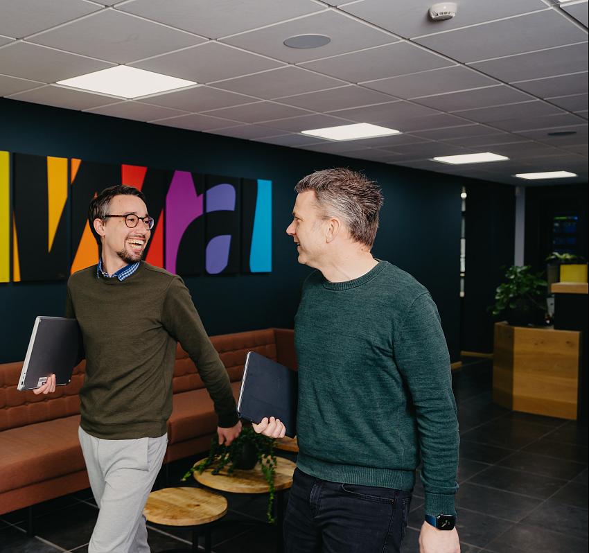 Twee collega's lopen lachend door een moderne kantoorruimte met kleurrijke kunst aan de muur en een laptop in de hand.